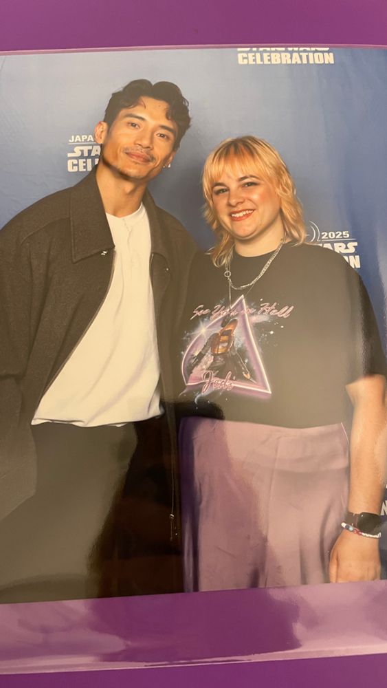 SWCJ 2025 photo op with Manny Jacinto of The Acolyte. He’s wearing black trousers, white shirt with a large, long brown coat. Me, the fan, in silk purple skirt, black t-shirt of Mae Aniseya within a neon purple triangle that says “see you in hell Jedi”