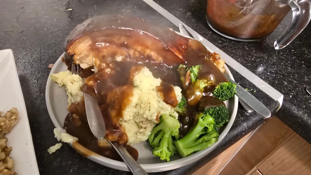 A roast dinner of Slow cooked Pork crackling joint, stuffing, creamy mash potatos, steamed broccoli covered in pork gravy on a round, gray slate plate on top of a black kitchen counter top