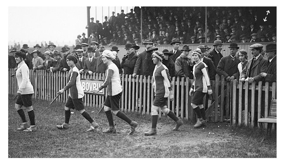 A black and white photograph of five women football players from the period of WWI in Britain walk onto a pitch, with a fence separating them from a large crowd.