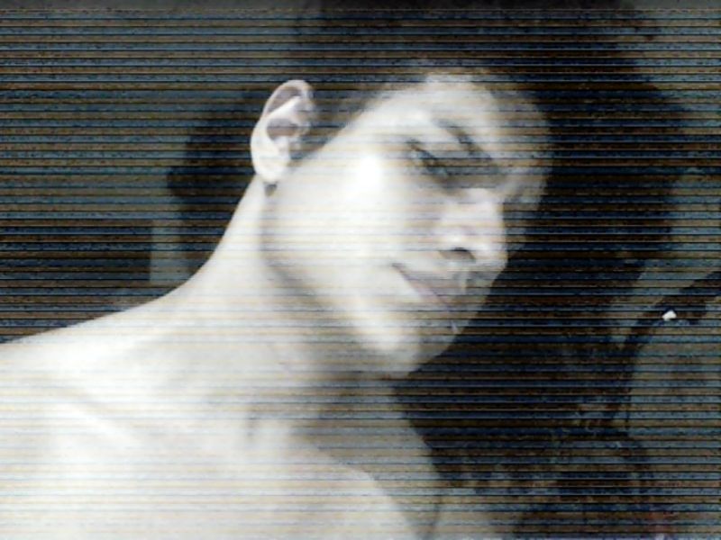 homem descamisado, de cabelos cacheados pretos e aparentemente branco olha inclinado para ao lado com expressão de suspeição na direção da câmera, a qual apresenta uma resolução fudida fruto da sua natureza vga 480p e a imagem é em preto e branco. guidão de bicicleta e vão de porta podem ser vistos ao fundo