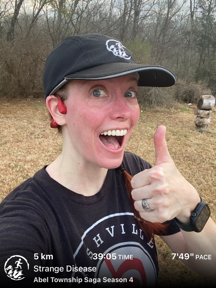 Fair skinned girl with black shirt and black hat and a red face from running giving a thumbs up and smile 