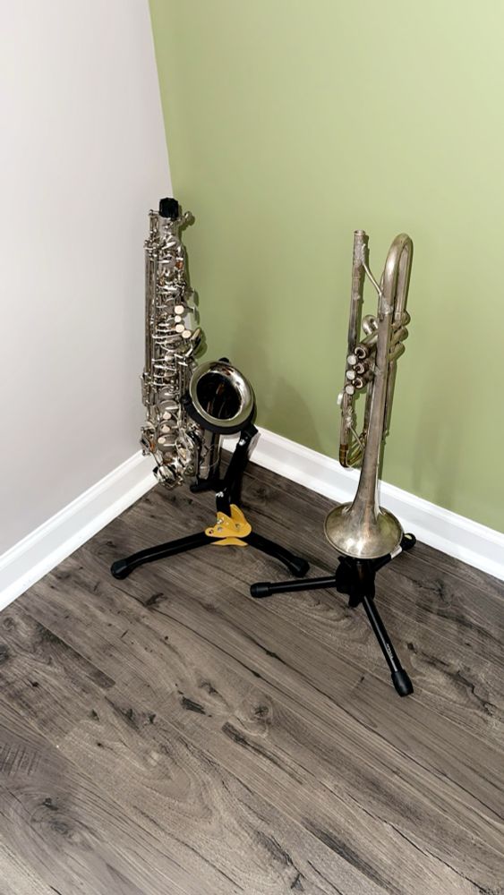 Picture of silver alto sax on a stand next to a silver trumpet, also on a stand.