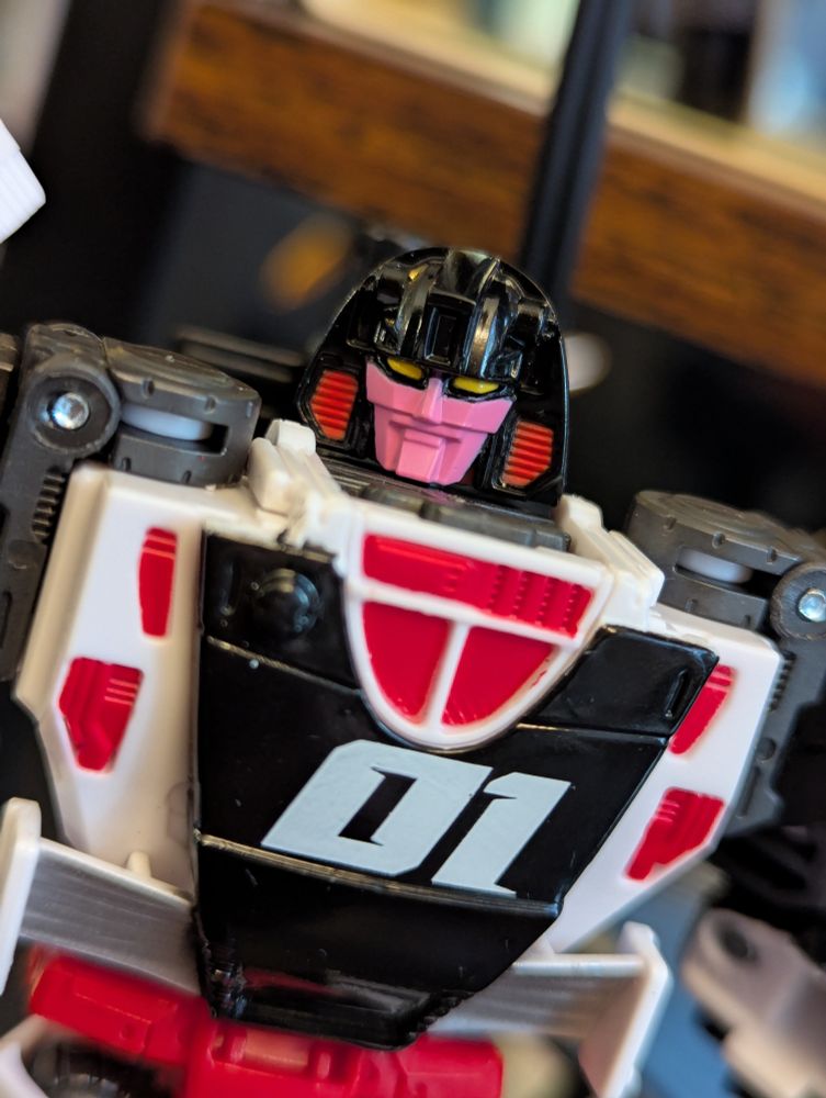 A close up of Crasher's head sculpt. It's a striking pink face surrounded by a glossy black helmet with red accented vents on either side. She has piercing yellow eyes and a square sculpted chin with a square sculpted lip that stops under a wider squared off upper lip which gives her a permanent grin. A very classic looking robotic face.