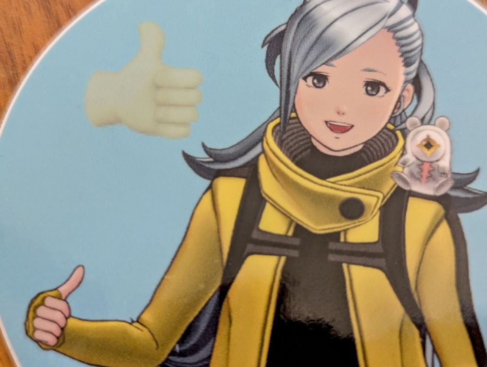 A sticker of Mizuki from AI: THE SOMNIUM FILES giving a thumbs up with a faded icon of the thumbs up emoji hovering near her head and Aiba in her eyeball form sitting on her shoulder. The background is a plain light blue.