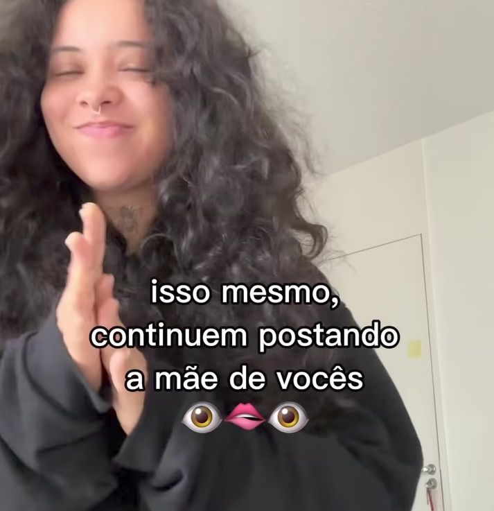 Print de um vídeo do insta onde uma moça bonita de cabelo ondulado solto, casaco preto e sorriso malicioso esfrega as mãos com más intenções. No centro, está escrito "isso mesmo, continuem postando a mãe de vocês" emoji mordendo o lábio 