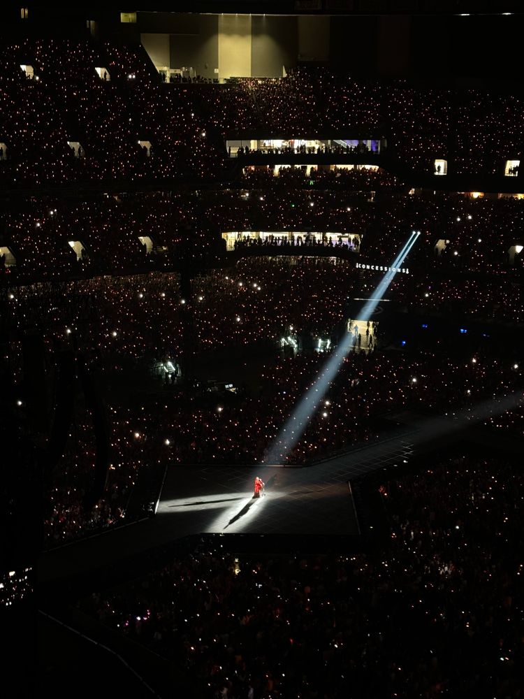 A zoomed out picture of The Eras Tour during all too well. Taylor is standing in the middle of the stage. The lights hit her and make an X on the stage. 