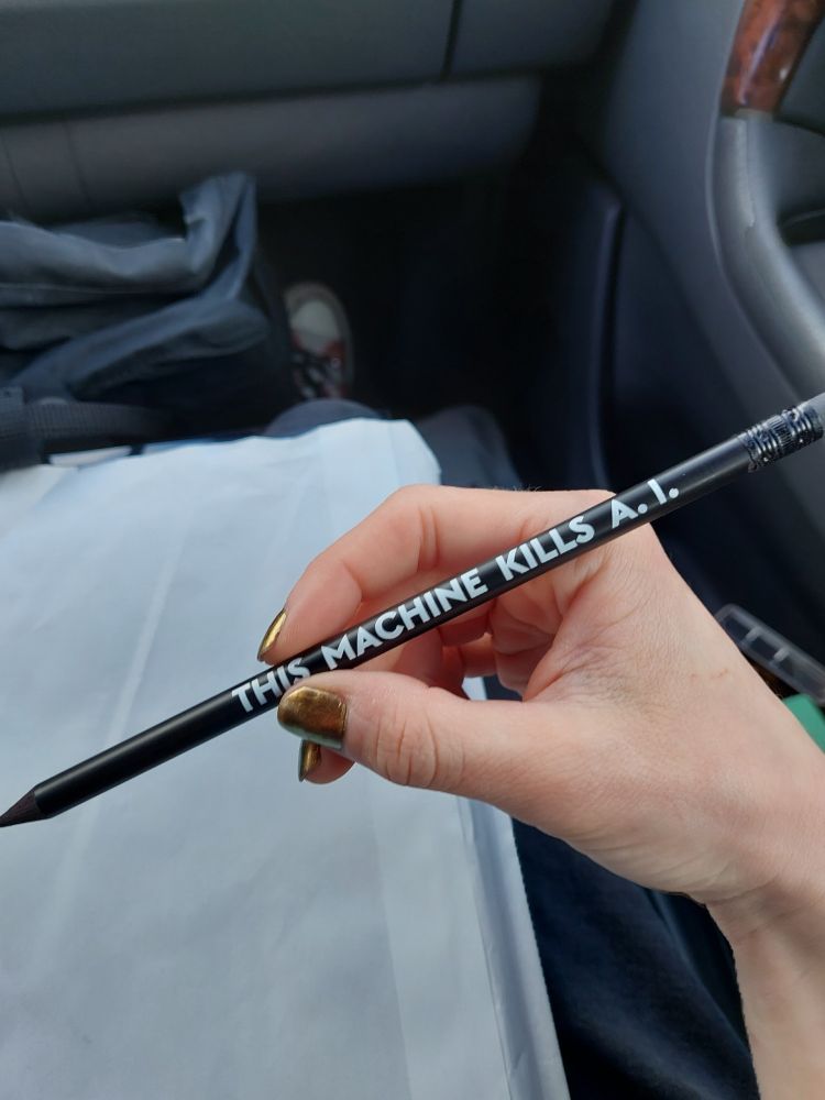 my hand holding a black wooden pencil  that reads "THIS MACHINE KILLS A. I." in white font