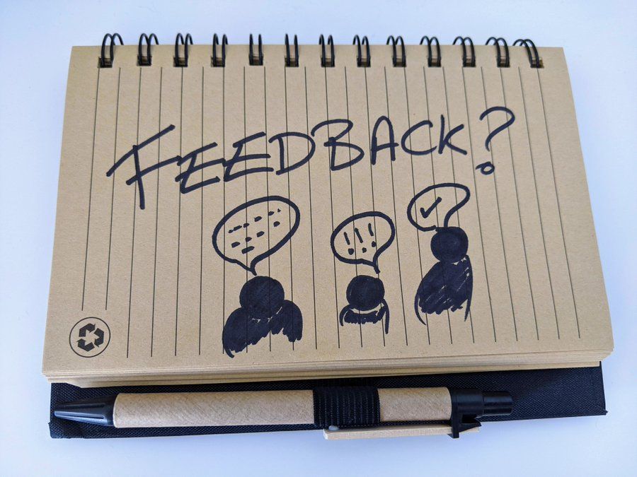 A spiral-bound notebook with lined pages is open, displaying a hand-drawn illustration. The word 'FEEDBACK?' is written in large, bold letters at the top. Below it are three simple figures with speech bubbles above their heads. The first figure's speech bubble contains dots, the second contains exclamation marks, and the third contains a checkmark. A pen is attached to the notebook at the bottom left corner.