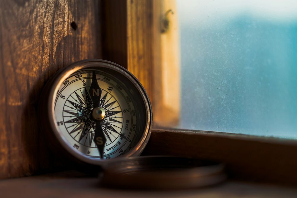 A vintage compass with a metallic casing, resting on a wooden surface near a window. The compass face shows cardinal directions with an ornate central design. In the background, a wooden window frame and frosted glass obscure the view outside.