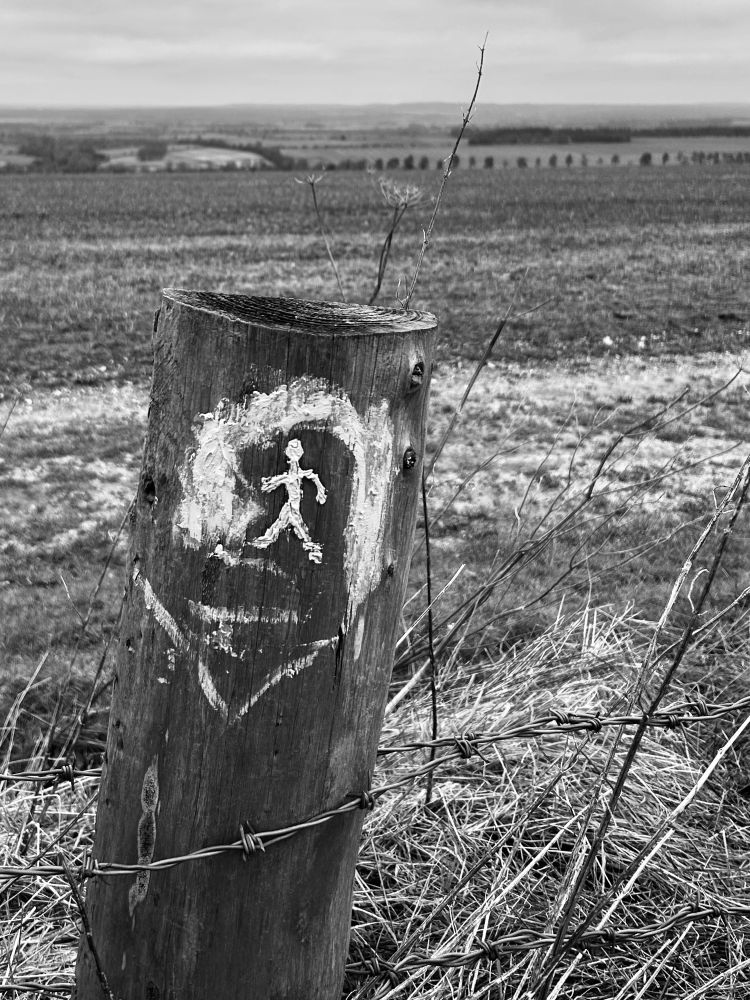 Black and white photo showing a small painting in chalk of a stick man on a fence post along the ridgeway with oxfordshire landscape behind.
