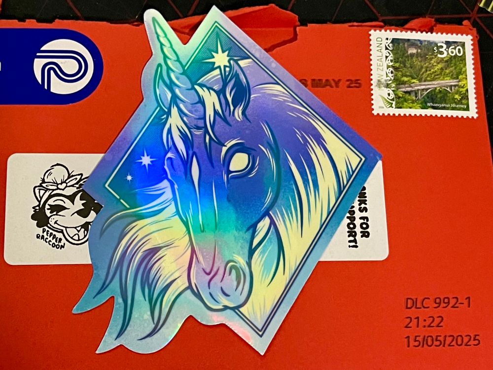 A shiny blue unicorn diamond sticker sitting on top of a red envelope sent by @pepperrqccoon.com from New Zealand. 