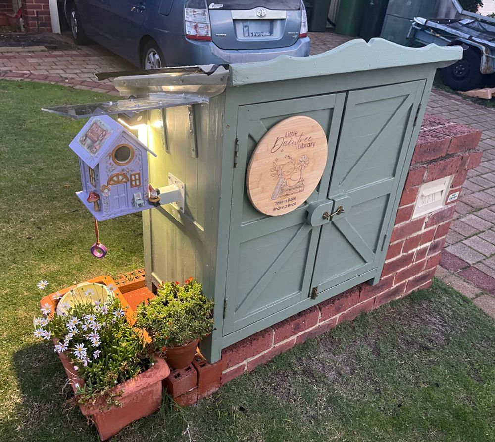 A big eucalypt-green Little Street Library built next to a red brick letterbox with some flowers planted to the left under a small purple house attached to the side of the library. 