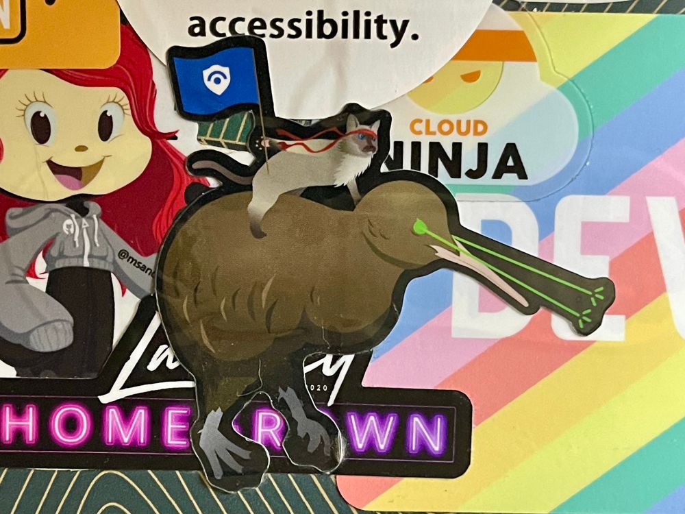 A sticker on a laptop of a ninja cat riding a giant kiwi with lasers coming out of its eyes. The ninja cat is holding a Microsoft Sentinel flag. 