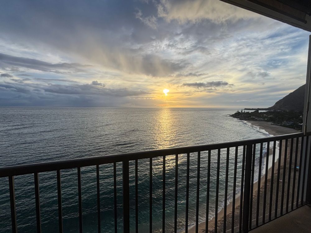 Photo of a beautiful sunset on the Pacific Ocean. The photo is taken from a high story balcony with views of a beach, abundant clouds, and a partially in frame mountain off to the right.