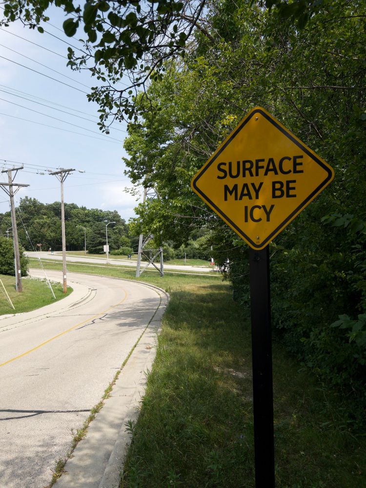 A yellow road sign says “surface may be icy” ahead of a curvy road surrounded with lots of green grass and foliage 