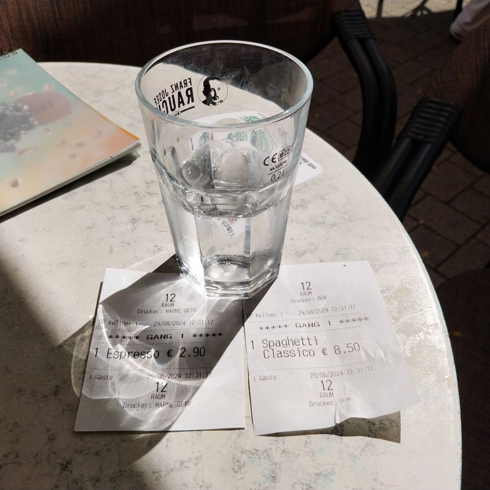 Ein runder Tisch, die Sonne scheint. Auf einer Terasse eines Straßencafe stehen Tische und Stühle, auf dem Tisch ein Glas Wasser und zwei Quittungen für einen Espresso und ein Spaghetti-Eis.