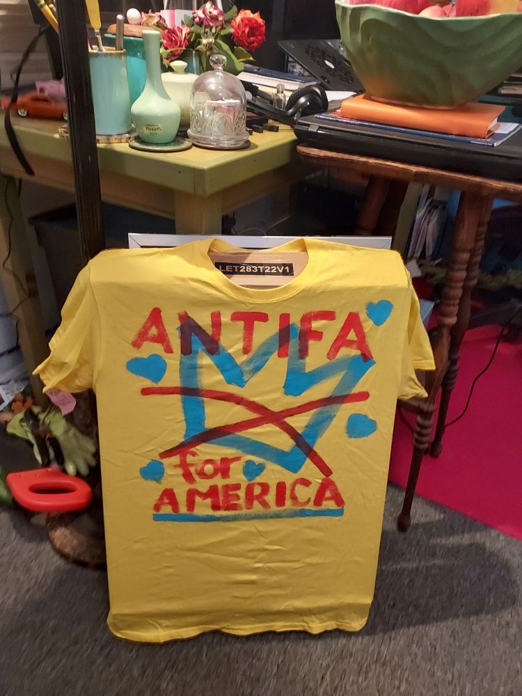 Yellow T-shirt that says ANTIFA for AMERICA with the NO KINGS crown with and X through it, and lil hearts.