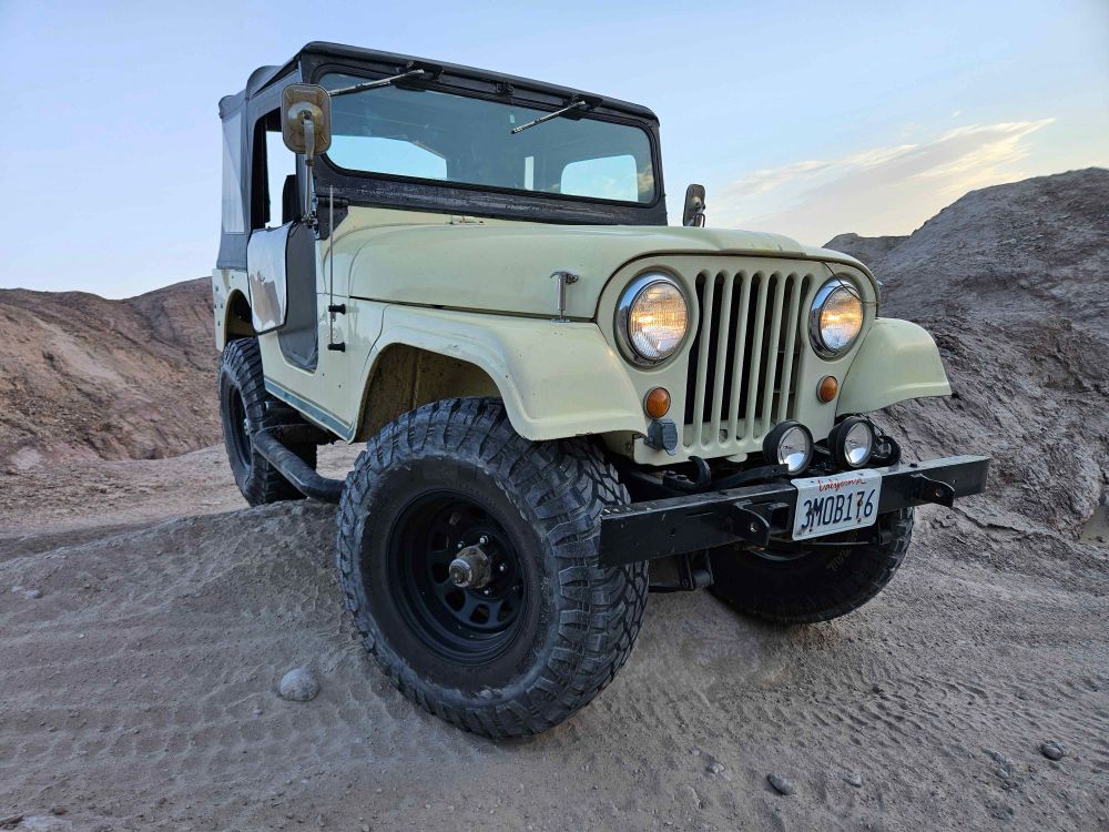 Tan dirt hills can be seen with a cream white jeep with black tires and a black top making a turn. 