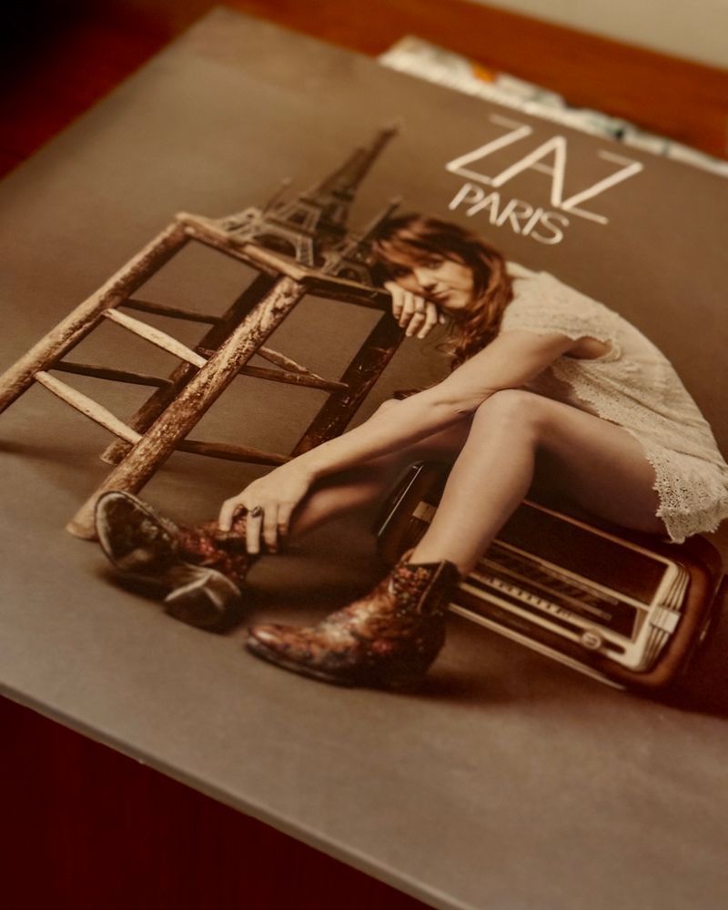 Das Albumcover von “ZAZ - Paris”, auf dem die Künstlerin auf einem alten Radio sitzt. Im Hintergrund steht ein kleiner Eiffelturm auf einem hölzernen Stuhl. Sie trägt ein weißes Spitzenkleid und auffällige Stiefeletten.