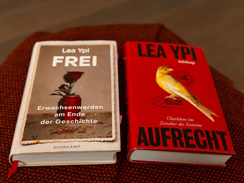 Zwei gebundene Bücher von Lea Ypi auf einem gewebten rotbraunen Untergrund: Links “FREI - Erwachsenwerden am Ende der Geschichte” (Suhrkamp Verlag) mit grauer Covergestaltung und Rosenillustration, rechts “AUFRECHT - Überleben im Zeitalter der Extreme” mit rotem Cover und einer goldgelben Kanarienvogel-Illustration.