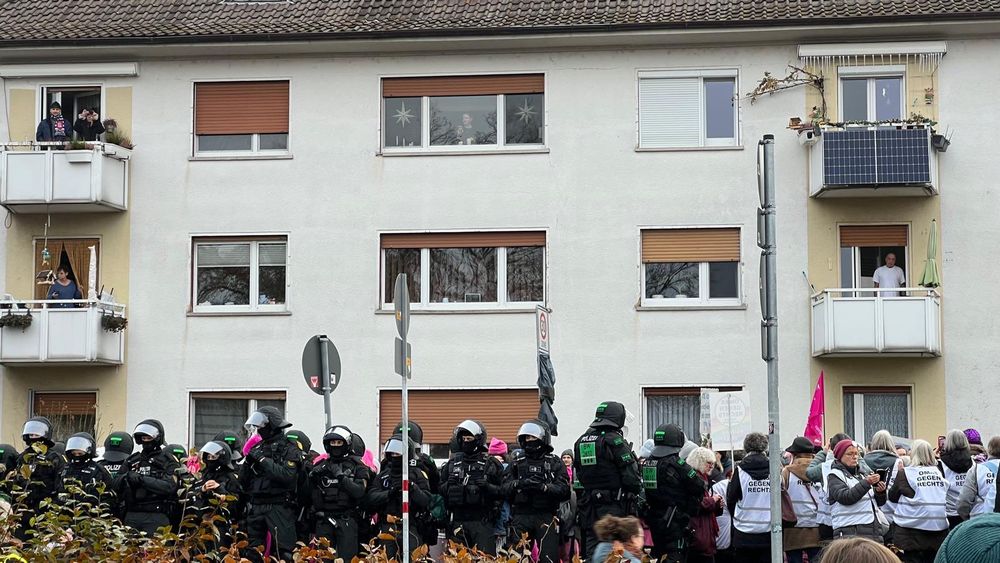 Wohnhausfassade mit mehreren Balkonen, davor eine dichte Reihe von behelmten und gepanzerten Polizeikräften, die einer Demonstration mit Transparenten und Schildern gegenüberstehen, während Anwohnende aus Fenstern und von Balkonen aus zusehen; rechts oben ein Balkon mit kleinen Solarpanels an der Brüstung.