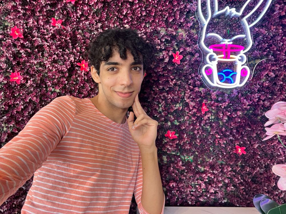 selfie of Madeline (me) wearing a peach shirt with white stripes. located in a boba place, there’s a purple floral background with an LED “neon-style” sign of a bunny with a cup of boba 
