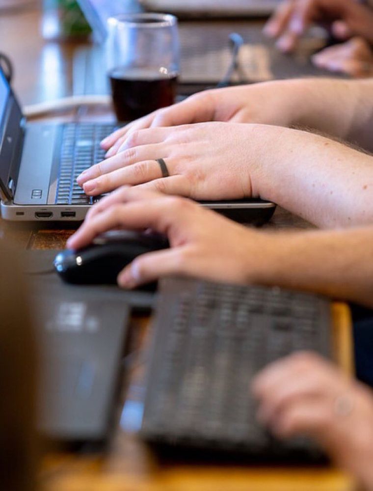 Close up image of three people's hands, individually working at laptops, one set of hands are typing, one is typing and using a mouse and the other using a trackpad on their laptop.
