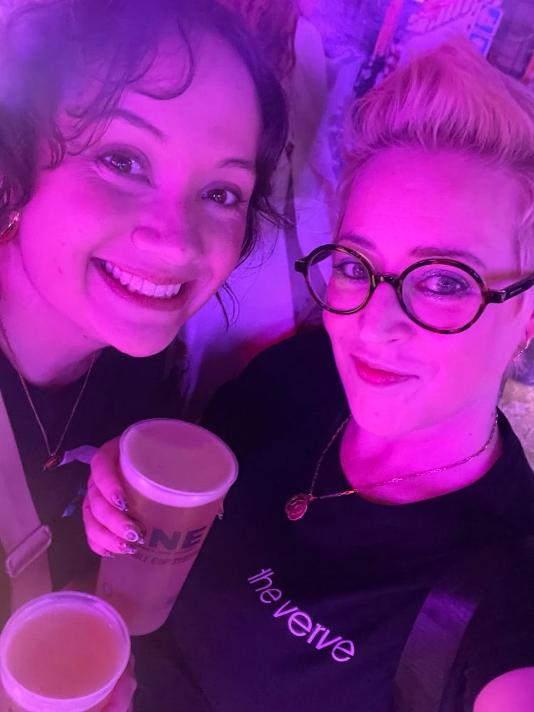 A selfie of Sarah and I in the Backstage Club. The low lighting is pink and lilac. We are both holding pints of Asahi lager and my T-shirt says “The Verve” in white letters on a black background 
