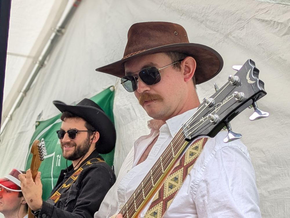 A close up of a guitarist and bass player performing with an out of shot band. They are wearing open shirts, sunglasses and cowboy hats. 