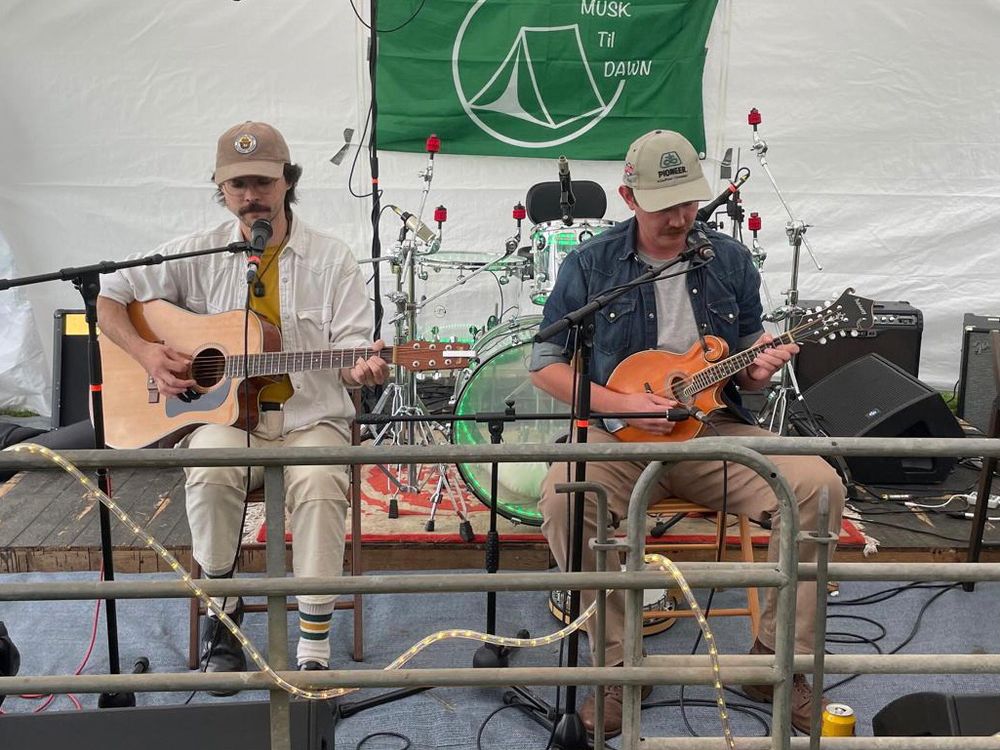 Two men performing on a stage in a festival marquee. One is playing acoustic guitar and the other is playing mandolin. Both have baseball caps and moustaches. 