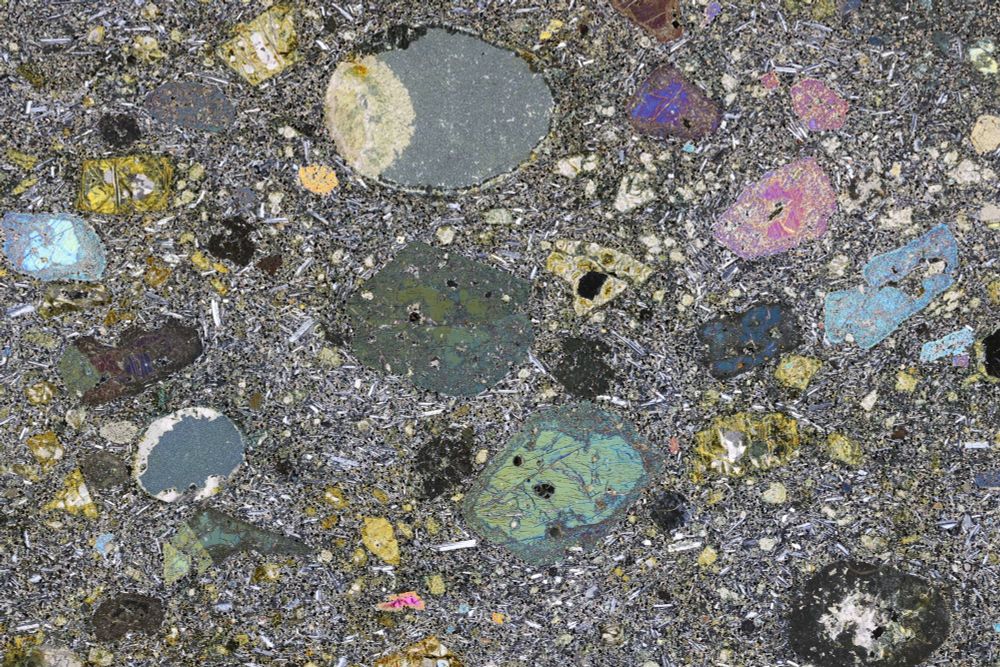 A photomicrograph of a basalt between crossed polars. It is full of large colourful clinopyroxenes, some round grey vesicles, and some yellowy grey altered olivines. The groundmass is full of little plagioclase feldspars and iron oxides.