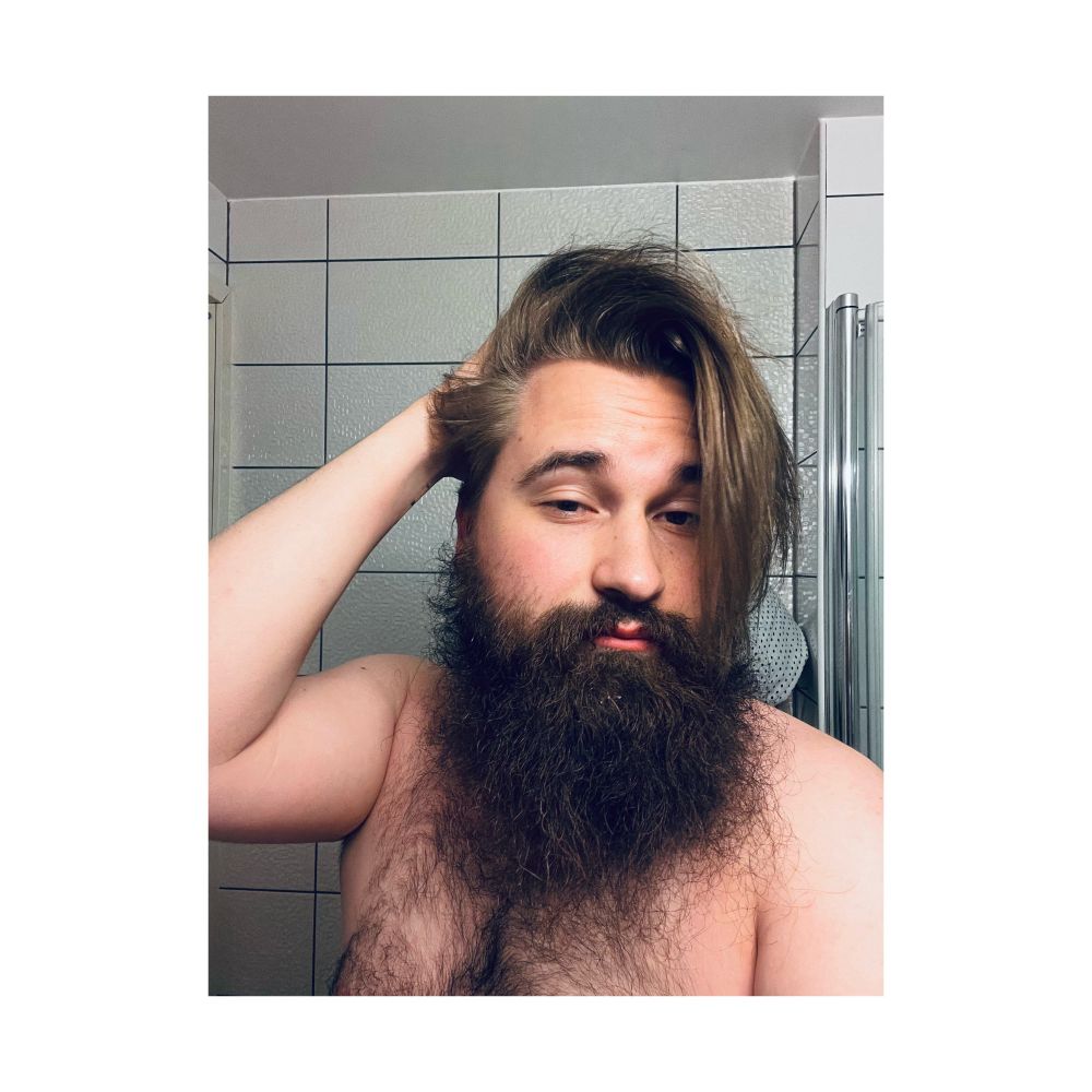 Nonbinary person with dark brown beard and light brown medium long hair posing in bathroom mirror.