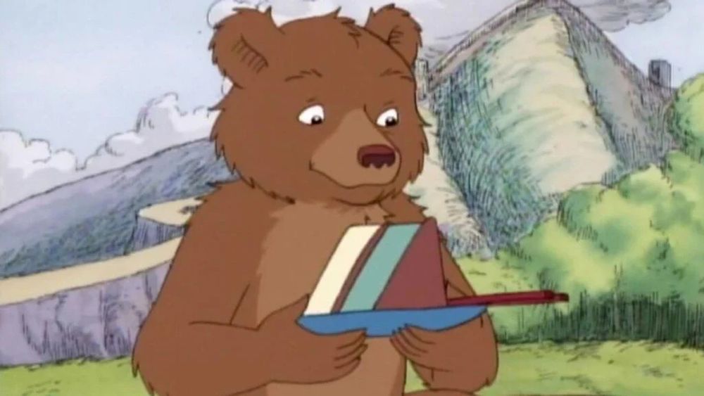 Screenshot from the show little bear. Little bear is holding a triangular object and the background is a cartoon forest. 