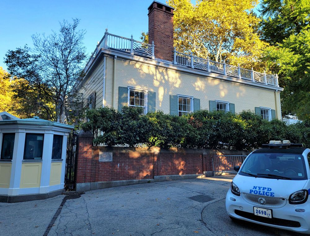 Daytime scene outside of an old historic mansion in a large city, surrounded by trees showing their fall colors; on the brick retaining wall surrounding the mansion is a plaque that reads "Gracie Mansion", for this is the official residence of the city's Mayor; also outside the mansion is a tiny "smart" police car and a small guard shack that matches the mansion's yellow paint scheme; the sky overhead is bright and blue. 