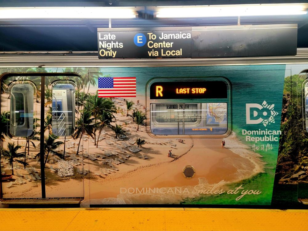 Daytime scene underground on a subway platform in a large city, facing a Queens bound R train at its last stop in Foreat Hills; hung above the platform is a black sign with white, Helvetiva lettering indicates that this track also serves the Jamaica-bound E local train; the subway car is completely wrapped in a photographic advertisement for Dominican Republic tourism, with aerial photos of its beautiful beaches stretching all across the car and, since the doors are closed, they become somewhat camouflaged by the beautiful imagery; at the bottom, it says "Dominicana smiles at you"; all along the edge of the platform are bright yellow safety treads that look like they were recently replaced or cleaned; the L.E.D. sign in the train car glows with the name of the line ("R") and "Last Stop" because this is as far as the train goes.