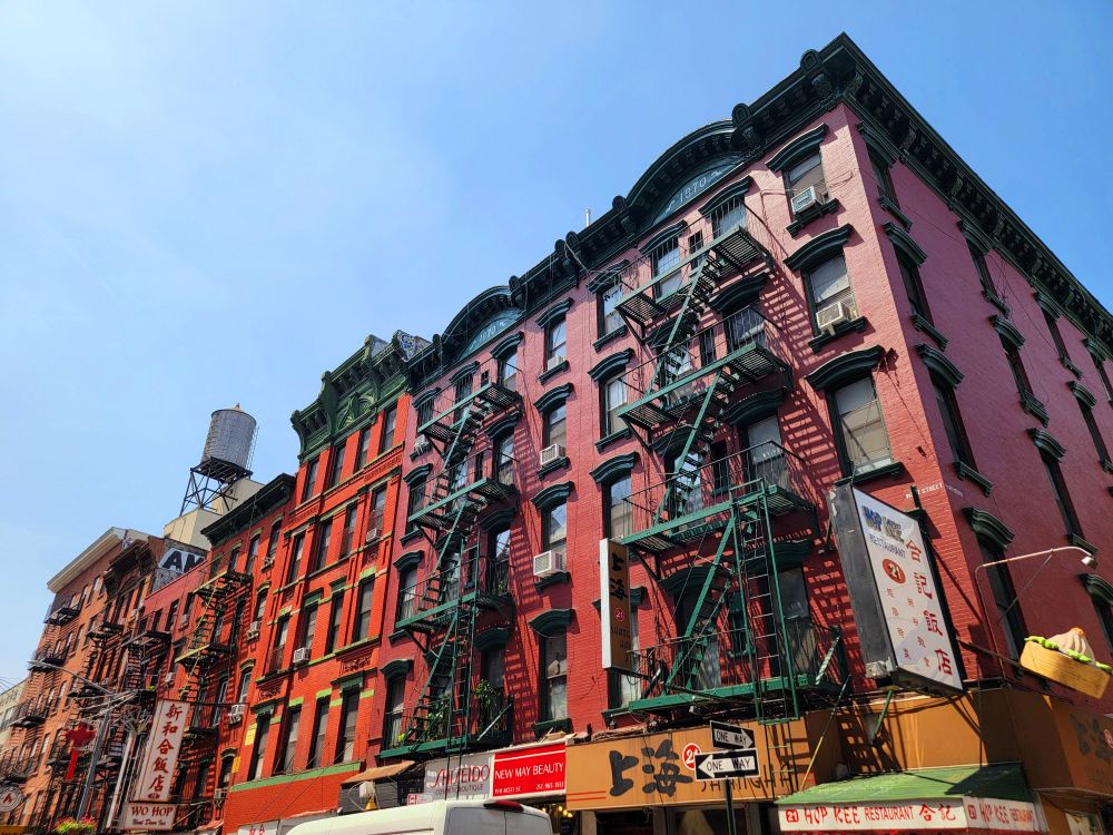Daytime view on a sunny day in a large city, looking upward at a group of very large, mid-rise, mixed-use brick buildings that were built in 1870; these bright red, historic buildings have beautifully-preserved green fire escapes, window transoms, and wall friezes; much of the detail of these buildings is very much Asian influenced, as this is in the heart of the city's Chinatown; in the spaces ar the street level of these buildings are a number of family-owned small businesses, including two of the most popular spots for Chinese cuisine - Hop Kee and Wo Hop, whose older signs are very simple since they do not need flashy signs to attract business but, rather, their legendary reputations - if you know, you know; atop the tallest of these buildings is a giant, cylindrical water tank; the clear blue sky in the distance shows less haze as it has had in recent days, as a light wind from the ocean is pushing back the wildfire smoke for the time being.