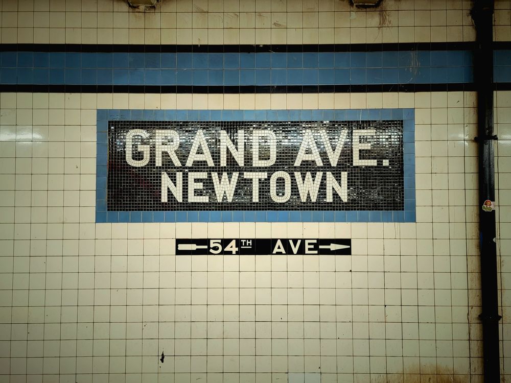 Below-ground view of the tiled wall of a platform at a subway station in a large city, featuring an inlaid mosaic feature reading "Grand Ave./Newtown" and a smaller inlaid tile reading "54th Ave" with arrows pointing in the direction of a staircase; attached to the tiled wall (to the right of the mosaic feature) is a black metal drainage pipe; the wall is definitely old and could use some cleaning, but the tilework is perfectly intact.