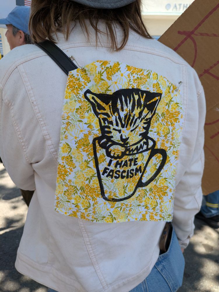 A woman at the Tesla takedown protest is wearing a floral fabric patch safety pinned to the back of her jacket. There is an adorable kitten sleeping in a cup printed on it, and the cup has "I Hate Fascism" written on it.