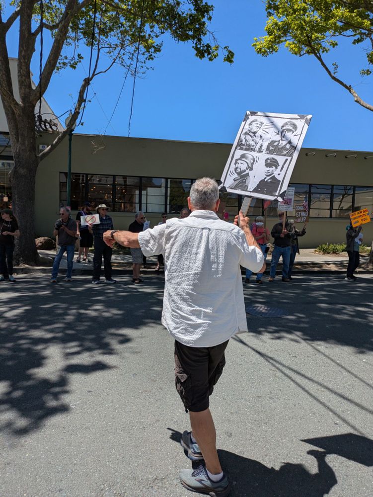 A man steps out into the street in front of the Tesla showroom to encourage passing cars to honk. He holds an amazing sign that features four black and white  photos of Trump, Musk, Vance, and Putin as various uniformed dictators from history.