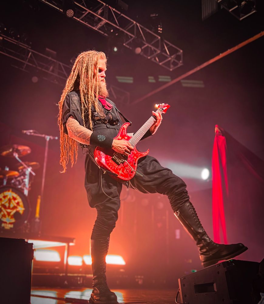 Picture taken by me of Jonas from the metal band Avatar is standing and wearing a black outfit with his leg propped up playing a red guitar.