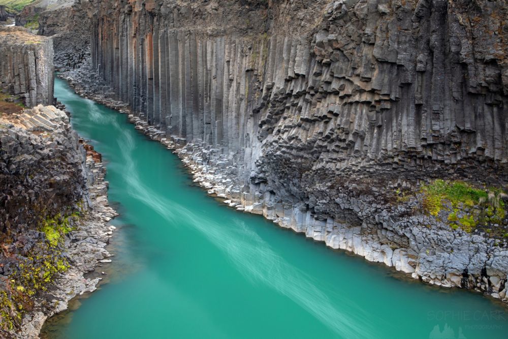 A green/turquoise river comes through a canyon from the top left of the photo and spreads outwards to the bottom. The river is surrounded by walls of basalt columns of different shapes, colours, sizes and directions. The exposure of the photo is slowed right down so there are some white patterns down the centre of the river. At normal speed these are ugly bits of dirty froth from the waterfall above!