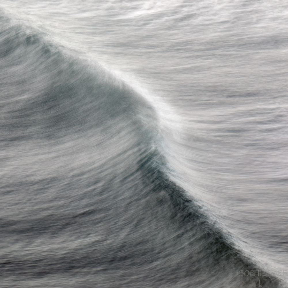A hand-held long exposure of a wave, with the swell going diagonally from top left to bottom right. The photo is square cropped and taken from above and the colours are mostly grey, with a touch of turquoise, white and darker grey where the wave is bulging before it breaks.