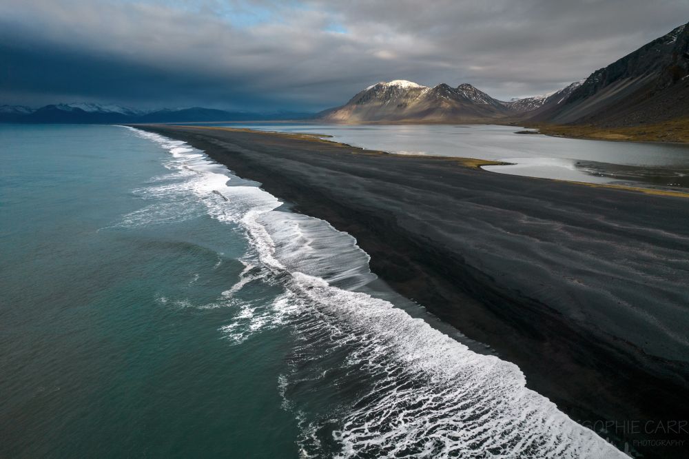 A drone photo of white waves crashing against a long black sand beach going from the bottom right of the shot diagonally towards the top left. The sea to the left is a green/turquoise. In the distance are some small mountains with dark clouds above, and a lagoon is seen behind the beach at the foot of a steep scree slope.
