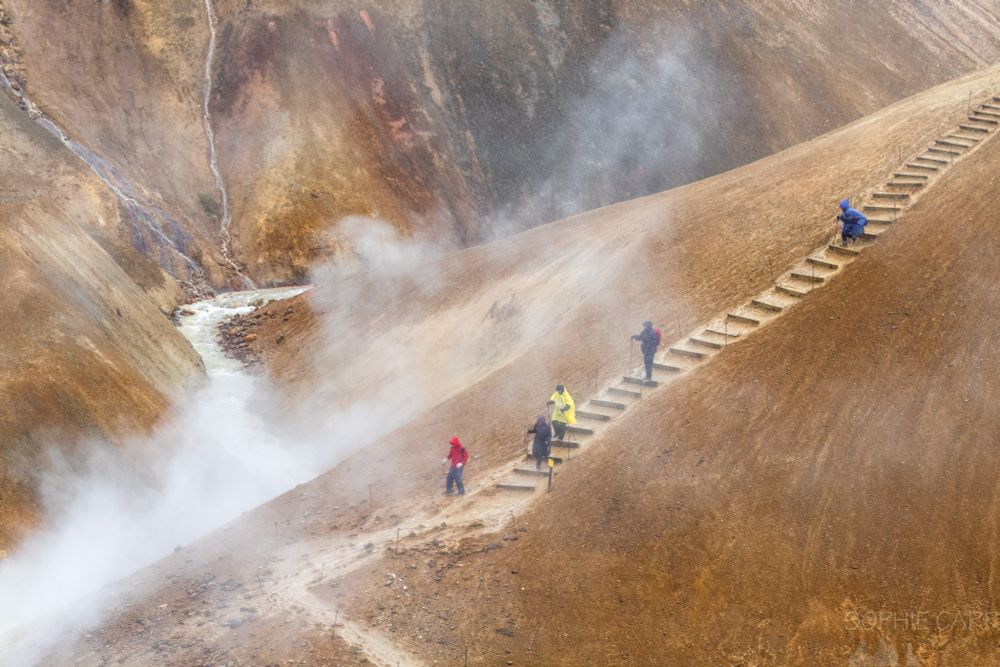 Five people wearing colourful rain jackets are seen hiking down some wooden steps on a steep orange hillside. Below some steam is rising from a small river which can be seen meandering off into the distance behind.