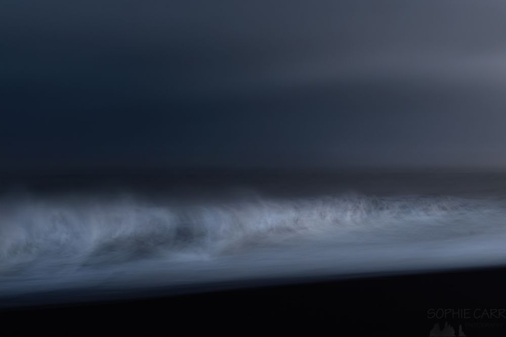 A dark photo of waves crashing against a black sand beach as the light fades after sunset. Using Intentional Camera Movement (ICM) to slow the movement of the waves and handheld - much easier than a tripod! Very dark ominous skies above the layer of light from the breaking waves.