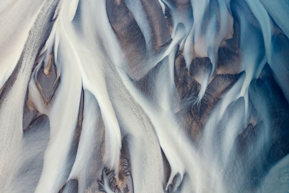 An abstract drone view looking directly down of a very meandering river estuary with lots of different braids. The water on the right hand side is very blue, whereas on the left it is almost white. There are some visible patches of black sand that looks a little golden in parts.