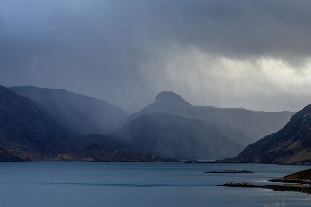 Visible rain in the distance above a loch. There are some mountains in the distance and clouds and some brightness above.