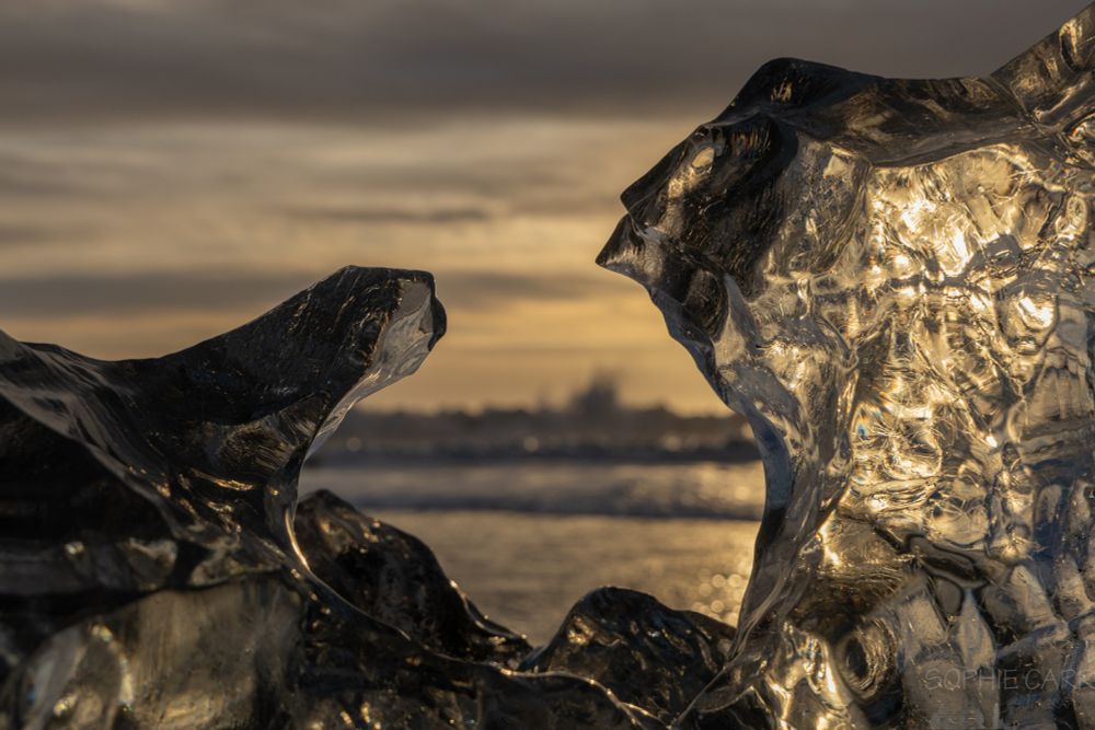 Close-up of an iceberg with a gap in the middle. The iceberg is golden and almost looks like two faces, facing each other. Between the sea can be seen, with a distant wave crashing. 