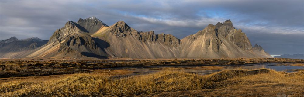Very wide panoramic shot of a stunning mountain with lots of peaks. It is golden hour and the light is shining on dead grasses in the foreground and midground, as well as on the peaks. There is some cloud and blue sky in the distance.