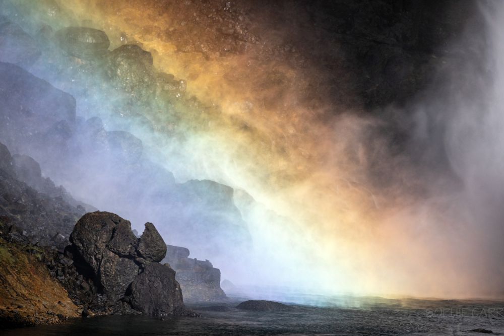 The photo is mostly of a rainbow - with some weird shaped basalt lava rocks at the bottom left and spray from a waterfall at the bottom right. The rocky bits look a bit like old troll heads, reclining & looking up at the view.